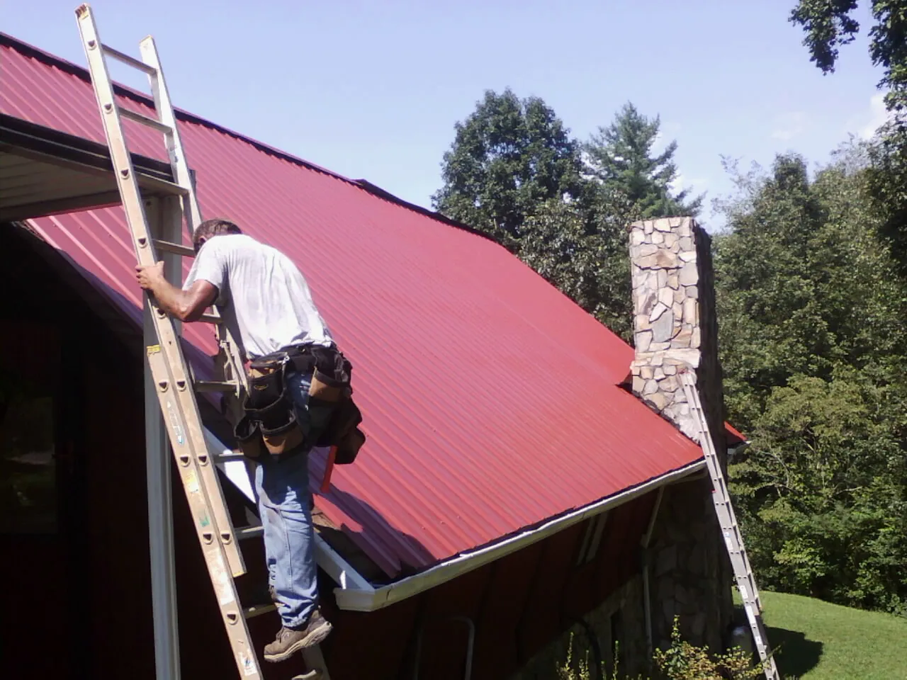 Roofer on a ladder beside a red metal roof and stone chimney — Soffit & Fascia Repair by Roofing Brothers in Alledonia